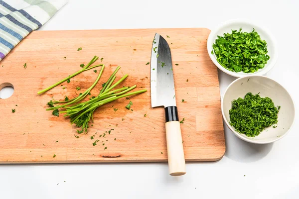 Dill sapları ve bir Santoku bıçağı mutfak tahtasının üzerine uzanır ve kase içinde dereotu ve maydanoz doğranır masanın üzerinde durur.