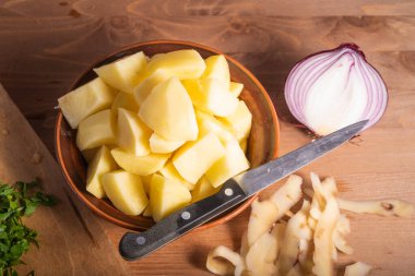 Vejetaryen yahnisi hazırlamak için gerekli malzemeler. Bir kasede soyulmuş ve doğranmış patates, yarım soğan, doğranmış yeşil soğan ve tahta bir masada patates kabuğu.