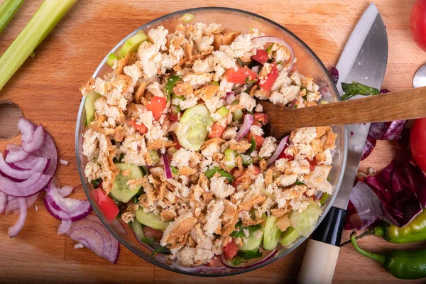 Cooking Italian Tuscan panzanella salad - bread salad, cucumbers, tomatoes, red onions, olive oil in a glass bowl with a wooden spoon, top view