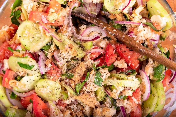 Cooking Italian Tuscan panzanella salad, mixed salad of bread, cucumbers, tomatoes, red onions, olive oil in a glass bowl, close-up, top view.