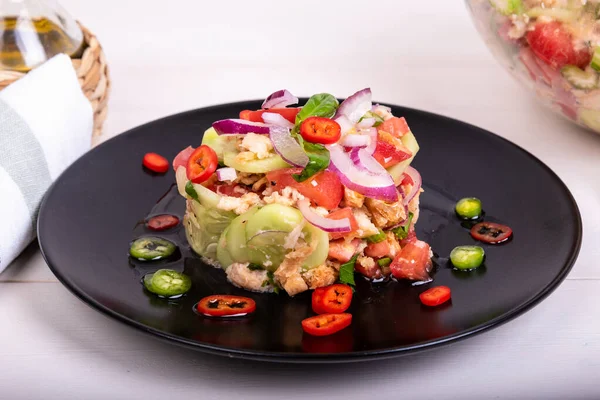 Italian tuscan salad panzanella, mixed salad of bread and vegetables on a black plate decorated with chilli peppers and a glass bowl with ready-made salad on a wooden light table, close-up