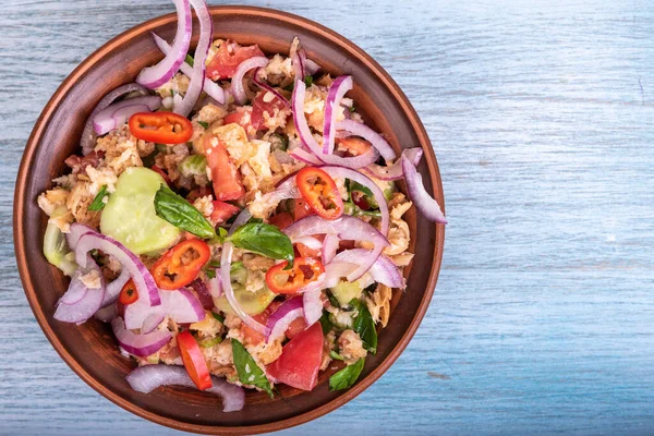 Italian tuscan vegetarian panzanella salad, mixed bread, tomato and vegetable salad in a clay bowl on rustic blue wooden table, top view, copy space