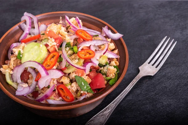 Italian tuscan vegetarian panzanella salad, mixed salad of bread, tomato and vegetables in a clay bowl on a black stone surface, top view, close-up