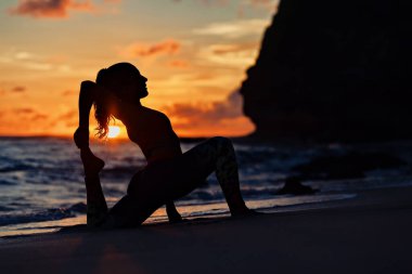 Gün batımında deniz sahilinde spor yapan genç bir kadın ayak hareketlerini sürdürmek için egzersiz yapıyor. Sağlıklı yaşam tarzı karanlık bir altyapı. Tropik ada yoga spa merkezindeki açık hava sporları, aile yaz tatili..
