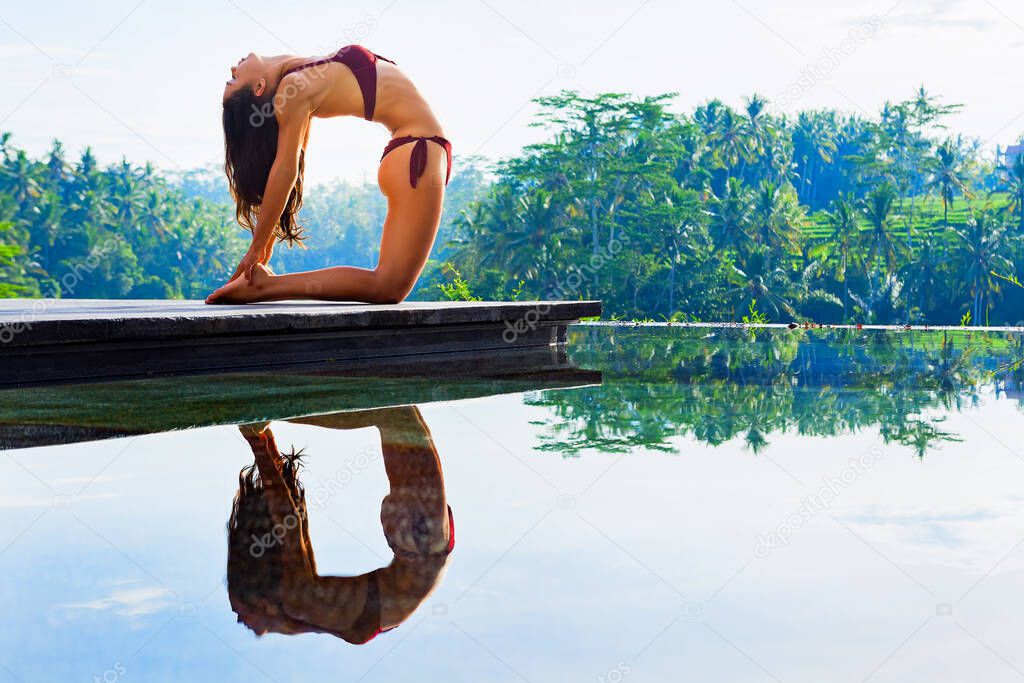 Stretching on sunrise background. Active woman in bikini practicing yoga at villa poolside to keep fit and health. Stand in ustrasana camel pose. Woman fitness, sport activity on summer family holiday