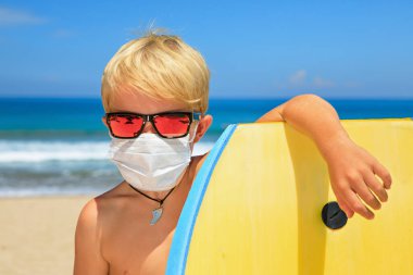 Güneş gözlüğü takan genç sörfçü, deniz kıyısında vücut tahtasıyla koruyucu maske takıyor. Yaz turları, Coronavirus COVID-19 salgını nedeniyle gemi seyahatleri iptal. Aile tatili için güvenli seyahat yerleri.