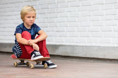Mutsuz ve üzgün görünen kaykaycı çocuk kaykay kursundan önce kaykaya oturuyor. Aktif aile hayatı, açık hava aktiviteleri, yaz tatillerinde çocuklar spor yapıyor..
