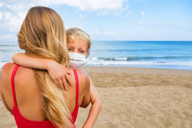 Anne deniz sahilinde çocuğa tıbbi maske taktı. Halka açık yerlerde bez surat takmak için yeni kurallar. Gemi gezisi iptal, Coronavirus COVID 19 turu. Aile tatili, 2020 yazında seyahat yaşam tarzı.