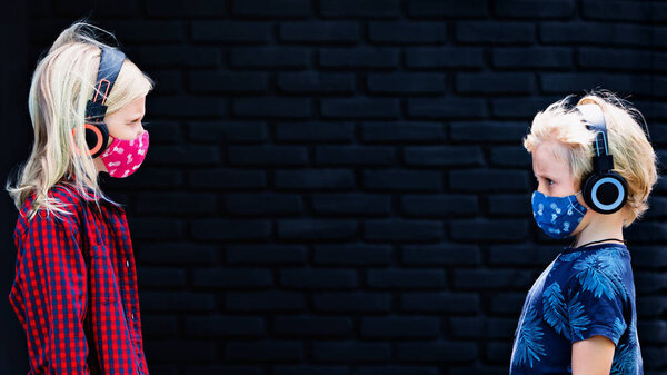 Смешные маленькие дети в наушниках, слушающие музыку, держат социальную дистанцию на улице. Дети в защитных масках для прогулок на улице из-за пандемии коронавируса Ковид-19.