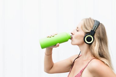 Beyaz arka plandaki genç sarışın kadın yeşil kullanılabilir şişeden taze kaynak suyu içiyor. Yaz şehrinde sağlıklı bir yaşam tarzı.