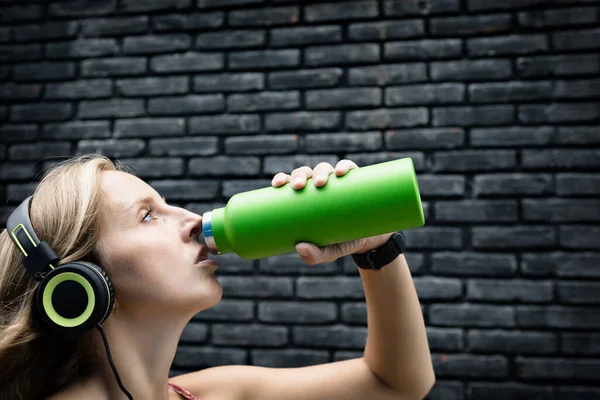 Siyah arka plandaki genç sarışın kadın yeşil kullanılabilir şişeden taze kaynak suyu içiyor. Yaz şehrinde sağlıklı bir yaşam tarzı.