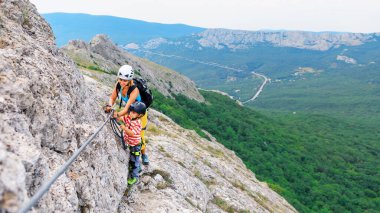 Genç anne, güvenlik ekipmanlı çocuk ferrata başlangıç rotasından tepeye tırmanıyor. Aile seyahati macerası, yürüyüş aktivitesi. Çocuklar yaz tatilinde doğayı keşfediyorlar. Hafta sonu yürüyüş turu