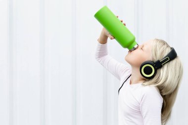 Beyaz arka plandaki genç sarışın kız yeşil kullanılabilir şişeden taze kaynak suyu içiyor. Yaz şehrinde sağlıklı bir yaşam tarzı.