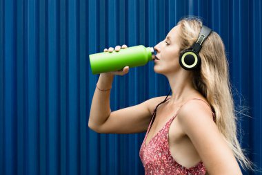 Mavi arka plandaki genç sarışın kadın yeşil kullanılabilir şişeden taze kaynak suyu içiyor. Yaz şehrinde sağlıklı bir yaşam tarzı.