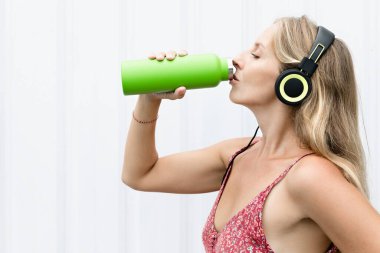 Beyaz arka plandaki genç sarışın kadın yeşil kullanılabilir şişeden taze kaynak suyu içiyor. Yaz şehrinde sağlıklı bir yaşam tarzı.
