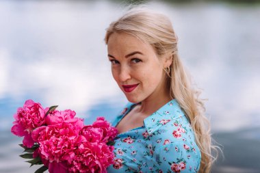 Elinde pembe şakayık demeti olan genç sarışın bir kadının portresi mavi elbiseli ve çiçekli şapkalı bir kız mavi suyun arka planında dinleniyor. Açık hava eğlencesi konsepti. Yaz tatili.