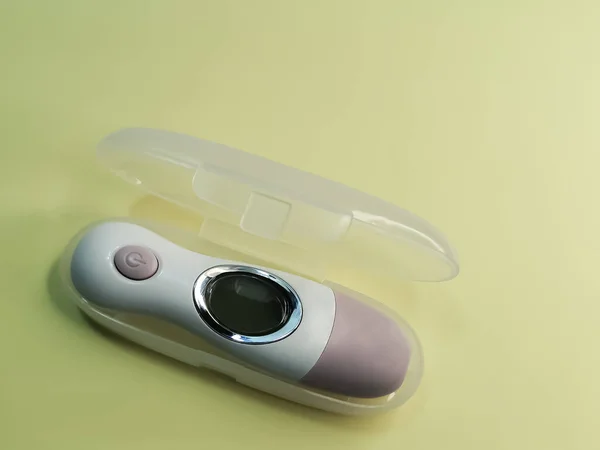 White electronic non-contact thermometer for measuring body temperature in children and adults on pale yellow background. The device is in individual transparent plastic case. Modern medical devices