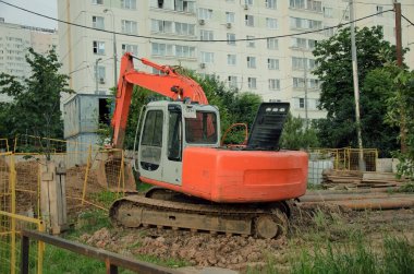 Moskova / Rusya - 05 / 07 / 2020: Bir evin arka planında kırmızı bir gövdeye sahip çalışamaz inşaat kazıcısı. Küçük kentsel inşaat sahalarında inşaat malzemeleri ve kanalizasyon onarımı.