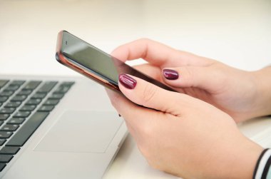 Parlak renkli tırnakları olan kadın eller akıllı telefonu dizüstü bilgisayarın arka planında tutuyor. Modern iletişimin sembolü internet ve mobil ağdır..