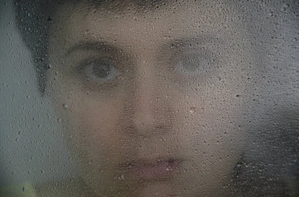 Portrait of a young brunette woman behind glass with rain drops. The concept of depression, blurring about the future, determination. Focus on drops on glass.
