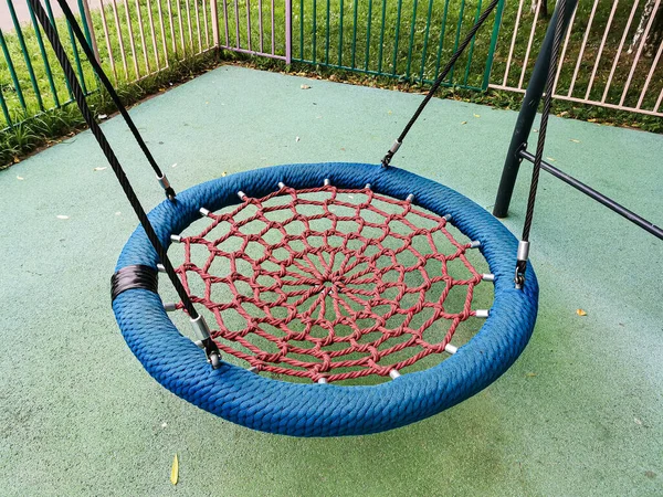 Kırmızı-mavi salıncakta asılı halatla örülmüş bir salıncak ve yerleşim alanındaki bir çocuk parkında hasır bir koltuk. Çocuk parkının yapay yumuşak bir örtüsü var..