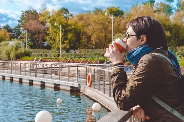 Young woman in profile drinking coffee, stands on the pier of city pond and looks at the water against the background of autumn park. A woman in autumn clothes with scarf wearing sun glasses