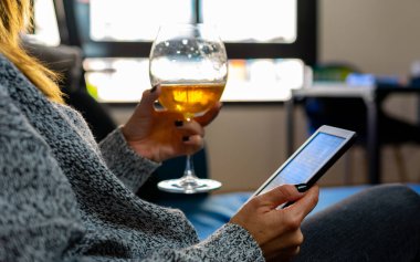 Tırnakları boyalı bir kadın, sağ eliyle ön planda bir e-kitap ve sol eliyle bir bardak bira tutuyor, evinde rahat ve sakince bir koltukta oturuyor, ışık evinin arkasındaki pencerelerden içeri giriyor.