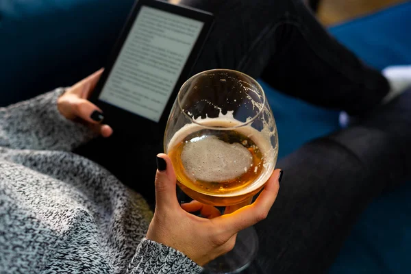 A woman with painted nails reads her e-book while drinking from a glass lying on an armchair