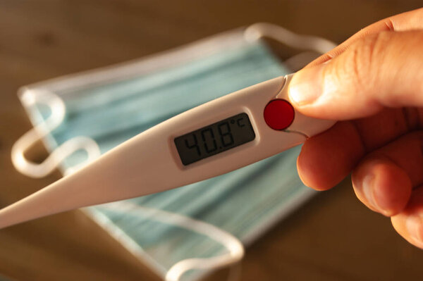 A hand holds a thermometer with degrees of fiber, and in the background you can see some surgical masks.