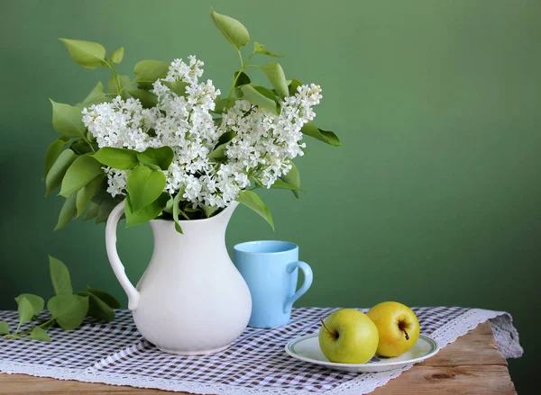 Blommande grenar av vit lila i en tillbringare och gula äpplen. — Stockfoto