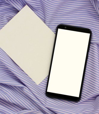 Beyaz ekranlı akıllı telefon ve çizgili mor kumaş üzerinde boş bir karton kart. Yukarıdan manzara. şablon, düzen, ürününüzü bağlamak için alan.