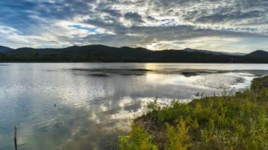 Bulutlu gökyüzü ve dağın altındaki dağ manzarası gündüzleri barajdaki suyu yansıtır.