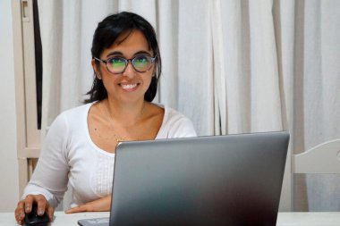 Evde bilgisayarla çalışan, gülümseyen ve kameraya bakan bir kadın.