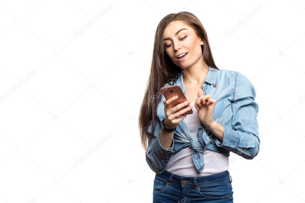 Portrait of a young brunette woman looking at the smartphone with surprised expression on her face against white background, isolated. Lifestyle, people and technology concept
