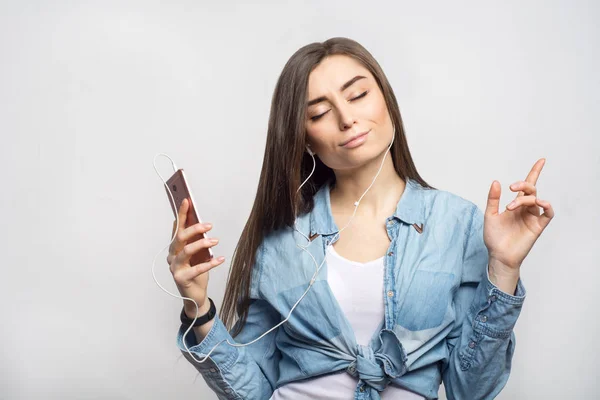 Retrato de una joven morena escuchando la música y bailando con un smartphone en las manos sobre fondo blanco, aislada. Estilo de vida, personas y concepto de tecnología — Foto de Stock