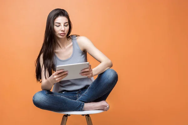 Retrato de una joven hermosa con una tableta en las manos sentada en la silla sobre fondo naranja. Estilo de vida, personas y concepto de tecnología — Foto de Stock