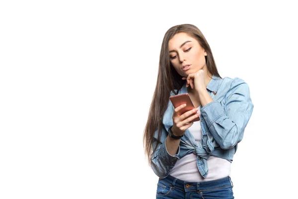 Retrato de uma jovem morena olhando para o smartphone em suas mãos com hesitação e miséria, pensando — Fotografia de Stock