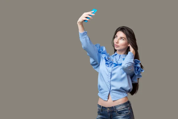 Портрет юной брюнетки в синей рубашке и джинсах, делающей селфи с помощью синего телефона, изолированного на сером фоне . — стоковое фото