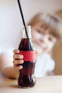 Gomel / Beyaz Rusya - 25 Temmuz 2018: güzel bir gülümseyen küçük sarışın kız coca-cola bir cam şişe bir masada bir borudan suni yeşil çim yeşil duvar tarafından içecekler