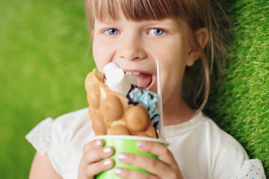 Güzel bir gülümseyen açık renkli kız bir yeşil kağıt bardak yeşil çimenlerin üzerinde bir gözleme ile lezzetli dondurma yiyor