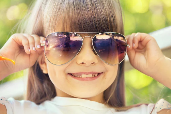 Güzel bir gülümseyen küçük ışık saçlı kız güneşli yaz Park'ta güneş gözlüğü takıyor bir köprü üzerinde yürür.