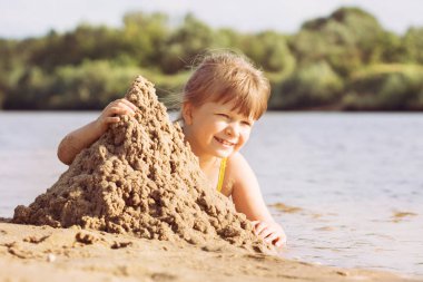 küçük sarışın neşeli kız bir mayo yaz aylarında güneşli sıcak havalarda nehir kıyısında kumdan kale oluşturur. portre