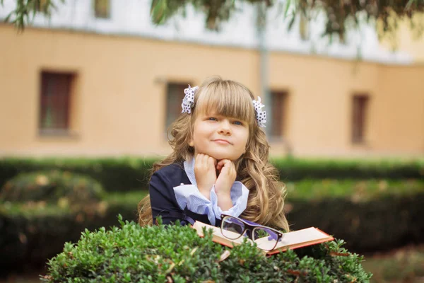 Bir küçük neşeli kız, mavi gözlü bir bantikami ve bir çanta ve bir kitap 1 Eylül ile okul üniformalı gözlük ile soluksuz saçlı birinci sınıf bir kadın parkta okul tarafından duruyor