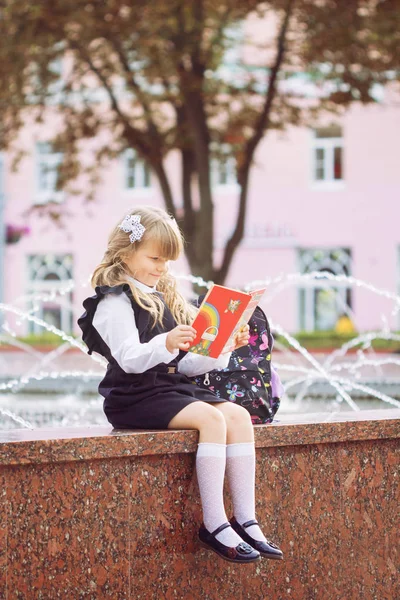一个小开朗的蓝眼睛的女孩 一个一流的女人 带着蝴蝶结和眼镜在学校制服与一个公文包和书在 9月1日 坐在学校公园附近的喷泉 — 图库照片