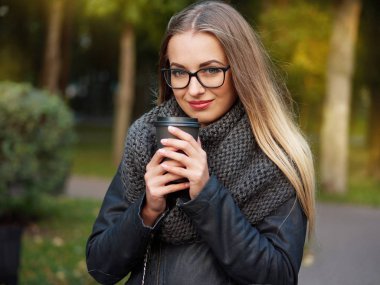 Güzel bir şık genç sarışın siyah makyajlı portresi bir deri ceket ve örme eşarp gözlük. Donmuş kız sonbahar soğuk parkta siyah bir kupa sıcak içecek içecekler