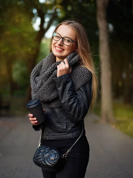 Güzel bir şık genç sarışın siyah deri ceket bardaklarda makyajlı ve örme eşarp bir çanta ile portre. Donmuş kız sonbahar soğuk parkta siyah bir kupa sıcak içecek içecekler