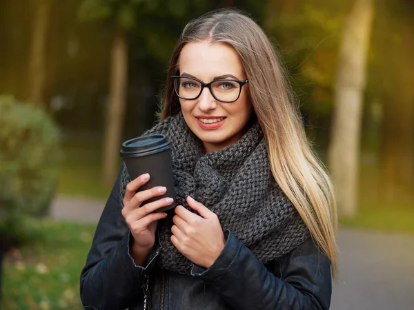 Portrait of a beautiful stylish young blonde with make-up in black glasses a leather jacket and a knitted scarf. Frozen girl drinks hot drink in a black mug in the autumn cold park