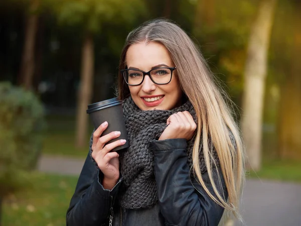 Güzel bir şık genç sarışın siyah makyajlı portresi bir deri ceket ve örme eşarp gözlük. Donmuş kız sonbahar soğuk parkta siyah bir kupa sıcak içecek içecekler