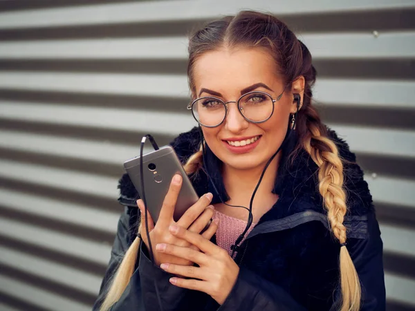 güzel şık genç bir kızla spikelets örgülü sarışın saç siyah koyun derisi ceketli ile kulaklık makyaj dinler bir çizgili demir amaç ona telefon üzerinden yuvarlak gözlüklü portresi