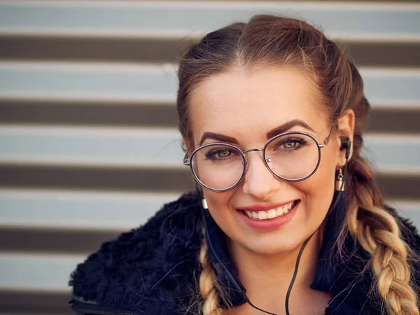 güzel şık genç bir kızla spikelets örgülü sarışın saç siyah koyun derisi ceketli ile kulaklık makyaj dinler bir çizgili demir amaç ona telefon üzerinden yuvarlak gözlüklü portresi